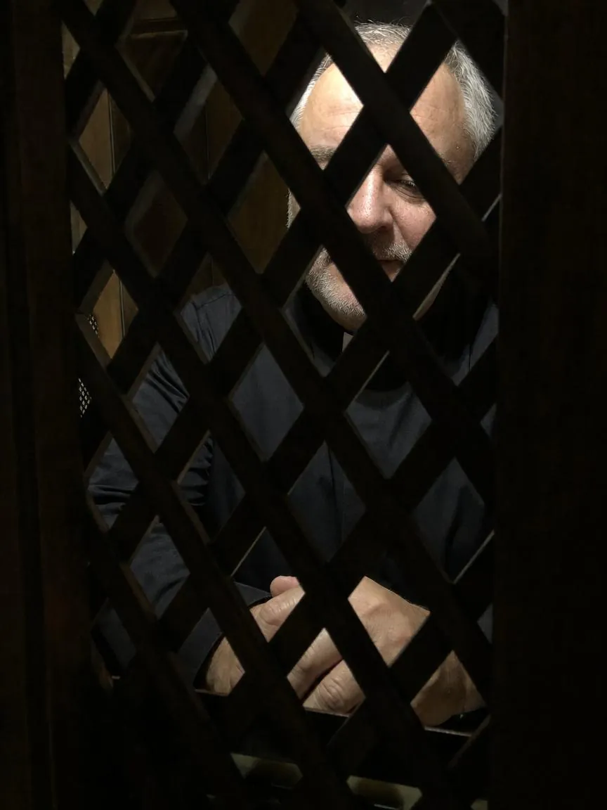 Fr. Marco inside a confessional, seen through the wooden grille