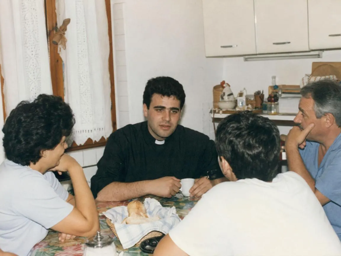 A young priest at a kitchen table with a family, early 2000s